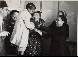 Helen Keller assists a young boy who is holding a chicken. They are surrounded by a group of people at a school for blind children. Egypt, 1952.