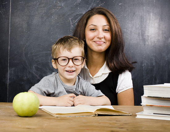 Little boy with teacher in classroom, sitting at table with books.