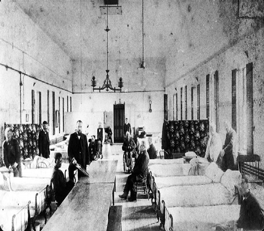 This is a view of a men's ward at Tewksbury Almshouse, 1898. The rectangular room houses two rows of neatly made beds and a center aisle with wooden tables. The room is very spare except for a chandelier. Patients sit and stand throughout the room, facing the camera, as does a gentleman holding what appears to be a clipboard. Image courtesy of the Public Health Museum, Tewksbury, Massachusetts.