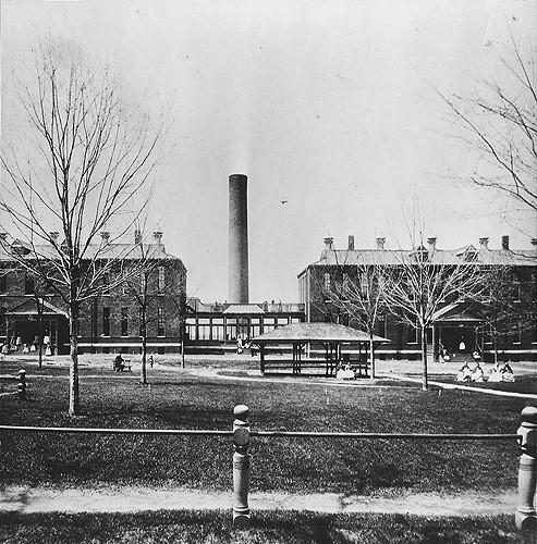 Exterior view of Tewksbury Almshouse, 1898. This view of the women's wards shows two brick buildings connected by a covered glass walkway. The chimney stack in the background is where the laundry was located. Patients are seen sitting in the yard in the foreground. Image courtesy of the Public Health Museum, Tewksbury, Massachusetts.