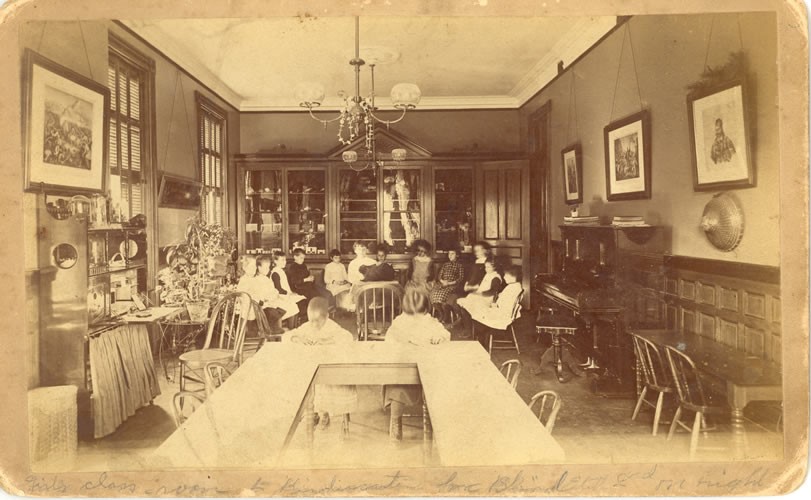 A classroom at the Perkins School for the Blind, circa 1890. In the background, students are seated in a circle around what is possibly a rabbit. Two students are seen working at a desk in the foreground. Two large windows create a bright atmosphere, enhanced by plants, a piano, and cabinets filled with equipment.