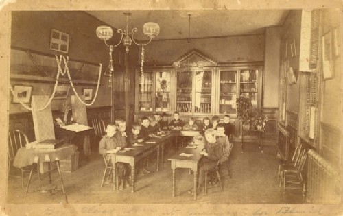 A classroom at the Perkins School for the Blind, circa 1890. The image is of the boys' kindergarten class. The pupils sit around tables placed in a U shape and appear to be working on a project. They are wearing either Victorian blazers and neckties or sailor suits, and they all have on lace-up boots. The room contains bookcases, plants, and pictures and is decorated with paper chains. There is no blackboard or teacher's desk.