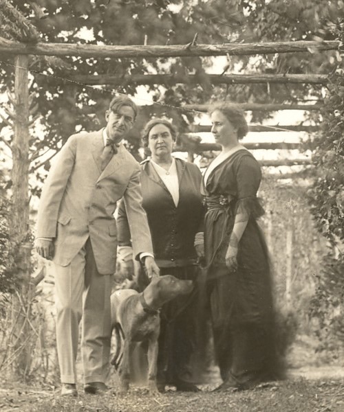 John, Anne, and Helen are shown standing with a dog in a garden, under a wooden structure covered in leaves, circa 1905. John is leaning slightly to his left, holding the dog's collar. He wears a light-colored suit with a white shirt and dark tie. Anne wears what appears to be two-piece dress with a long dark skirt, a matching hip-length jacket and a white blouse. Helen's dark dress is long, with a high waist and a white collar. The full, three-quarter length sleeves have a dark net ruffle at the elbow.