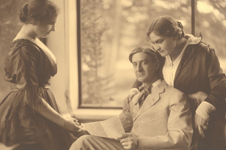 In this photograph taken at Wrentham, circa 1905, John is seated in three-quarter profile between Helen and Anne. Helen stands in front of him and Anne stands behind him. She leans against the back of his chair with her right hand on his shoulder. He is holding a manuscript and looking up at the camera while manually signing into Helen's hand. John wears a light-colored suit with a darker necktie that seems to be tied in a soft bow. Anne wears a dark, two-piece dress with a white blouse under the jacket. He