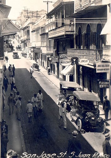 Photograph of San Jose Street, San Juan, Puerto Rico, circa 1915-1920s. Two Model A Ford type automobiles are parked in the sunshine on a commercial street. Wooden buildings and store-front signs line the street. Pedestrians walk down the middle of the street, some wearing round straw hats. Image courtesy of the Centro de Estudios Puertorriquenos.