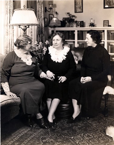 Photograph circa 1924 of (left to right) Anne Sullivan Macy, Helen Keller, and Polly Thomson seated in the living room of their home in Forest Hills, Queens, New York. Helen and Polly wear knee-length dresses. Helen's dress has a white ruffled collar, as does Anne's. Her dress is calf length. Helen's right hand is placed within Anne's left as if in the middle of manually signing to her teacher.