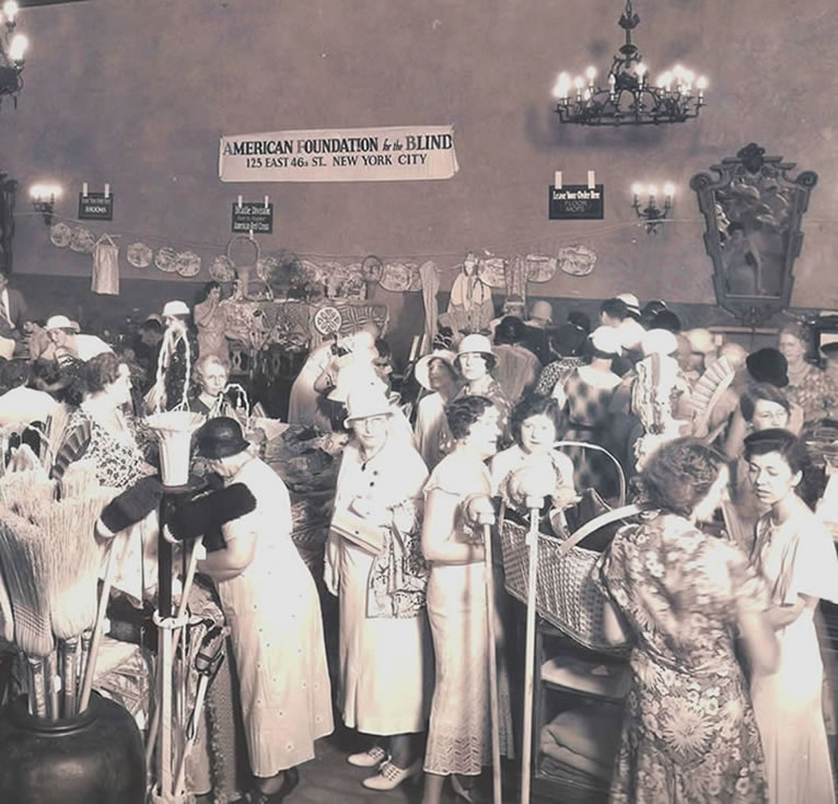 *Miami's First Educational Week for the Blind. Women in shin-length dresses mill around in a large hall where products made by blind workers are on display. Items include mops, brooms, a cradle, and sewn objects. A banner advertising the AFB-sponsored event is visible on the back wall, March 12, 1933. Verne O. Williams, photographer. American Foundation for the Blind Archives.* 