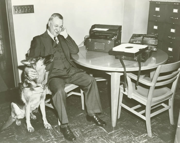 A blind gentleman with his guide dog sits at a table at the New York Public Library in New York City. He is listening to a record, which is being played on a Talking Book machine. Record sleeves and containers are laid out on the table. He rests his left arm on a table while his right arm is around the neck of his seated guide dog. The photograph is undated. Evelyn Strauss, photographer. Talking Book Archives, American Foundation for the Blind.* 