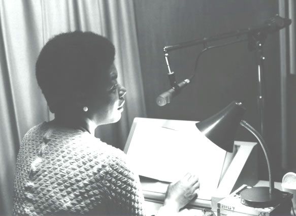 Author Maya Angelou is seen sitting in a recording booth at the American Foundation for the Blind, July 1970. She is photographed with her back to the camera reading from her book I Know Why the Caged Bird Sings. The book lies open and is illuminated by the table lamp. The lamplight illuminates the portion of her face that we can see. Talking Book Archives, American Foundation for the Blind.