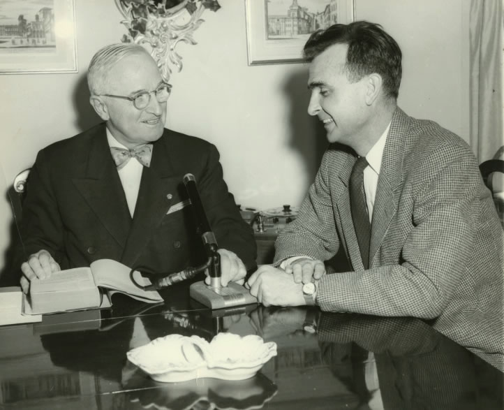 Former U.S. President Harry Truman (shown at left) is seated at a table with M. Robert Barnett, Executive Director of the American Foundation for the Blind. Truman, who is wearing a dark jacket and bow tie, is recording part of his book Year of Decision. Truman sits with one hand on his book and the other on a microphone stand. A reel tape recorder is just visible in the background between both men. New York City, February 3, 1956. World Wide Photos. Talking Book Archives, American Foundation for the Blind.