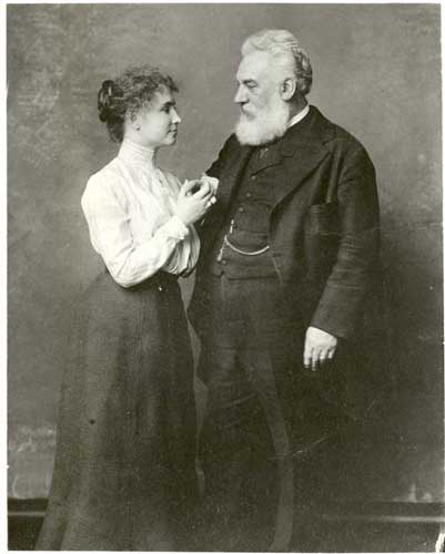 Helen Keller as a young woman standing with Alexander Graham Bell. He appears to be spelling into her hand. 