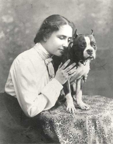 Helen Keller hugging a boxer-type dog who is standing on a small table. 