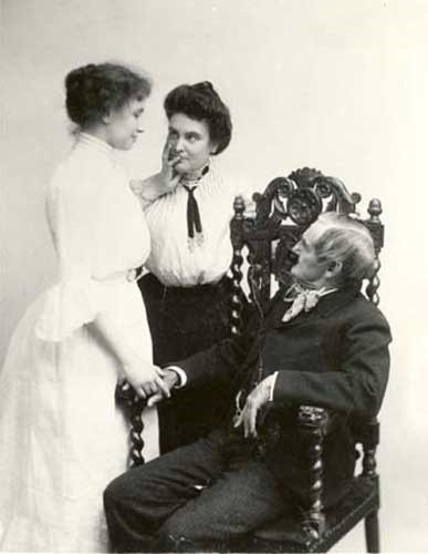 Helen Keller standing with Anne Sullivan, Helen's left hand on Miss Sullivan's lips and face. An older gentleman sits in a carved wooden chair gazing up at them and holding Helen's other hand. 