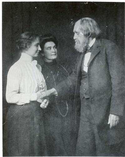 Helen Keller and Anne Sullivan with a tall bearded man. Helen is holding his hand.