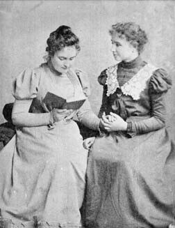 Helen Keller and Anne Sullivan seated. Miss Sullivan reads a book and spells into Helen's hand.