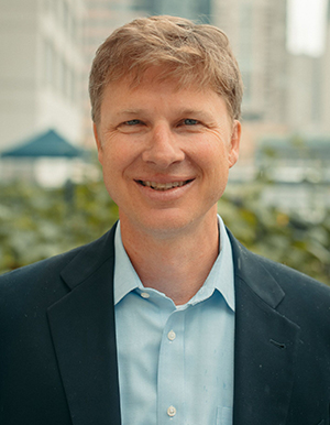 Headshot of Derek, a white male with short blonde hair, is smiling, wearing a blue coat and shirt. He is outside with trees and buildings far behind. 