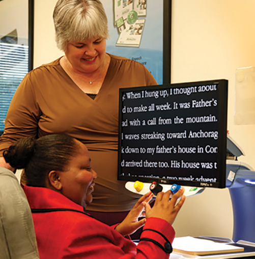 A woman stands next to a young woman sitting at a desk in front of a digital magnification screen.