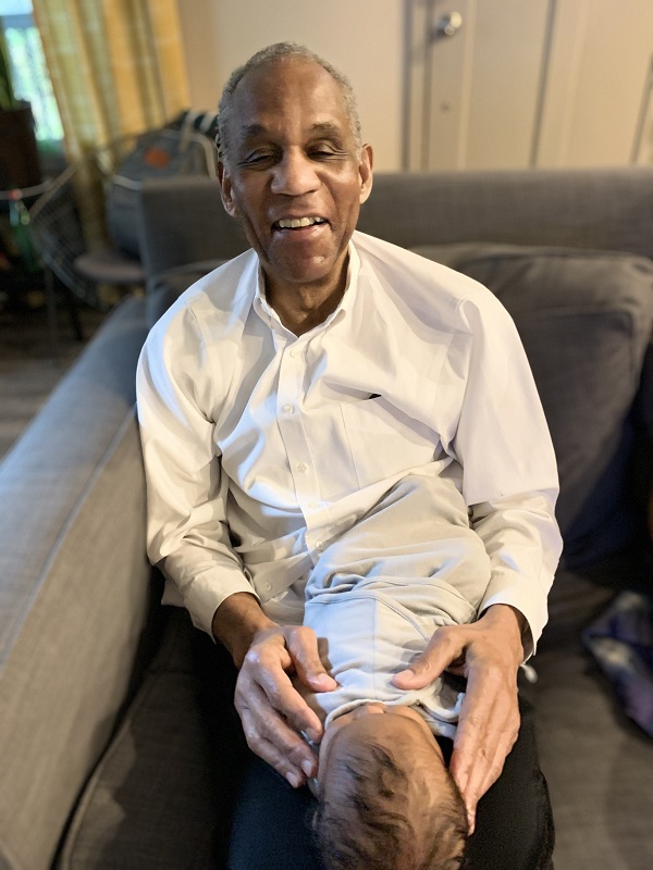 MacArthur Darby sits in a chair with his infant grandchild stretched out on his lap.