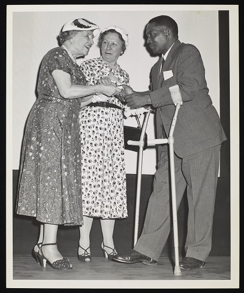 Standing (probably) at an award ceremony are left to right, Helen Keller, Polly Thomson and an unidentified man on crutches. They are pictured full-length and are probably standing on a stage. The man faces Keller as Thomson looks on. The man appears to be receiving a book from Keller. Everyone is smiling. The women wear floral print, calf-length dresses with short sleeves, hats and gloves. The man is wearing a suit and tie.