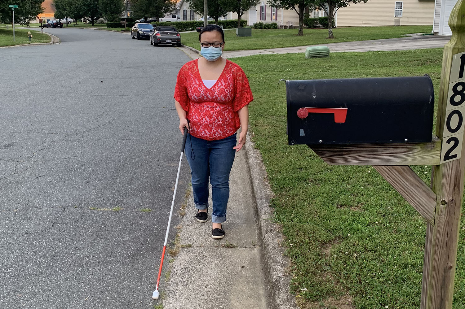 An Asian woman wearing a mask walks along a neighborhood street using a white cane.