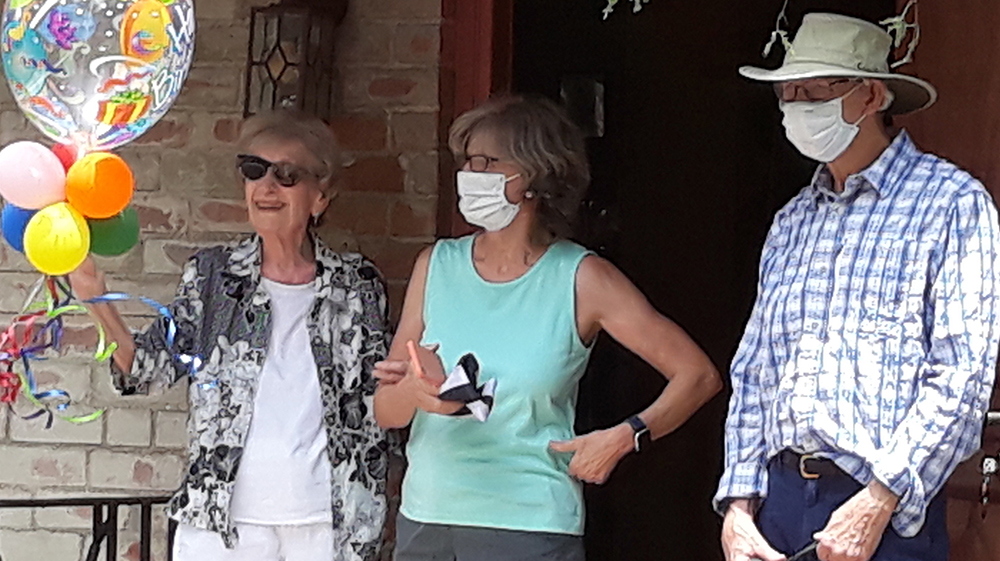 Three older White adults stand on a porch. One woman and the man wear masks and one woman does not. The woman without a mask is holding a bouquet of balloons. The man is holding the leash of a dog. 