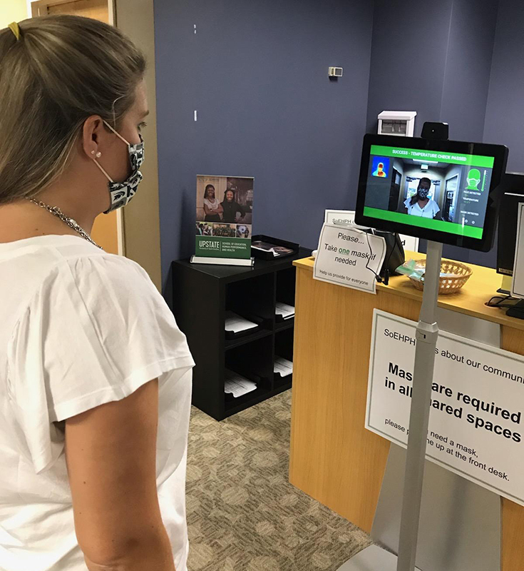 A young White woman wearing a mask is having her temperature checked by standing in front of a video monitor. Signage on the desk explains that masks are required.