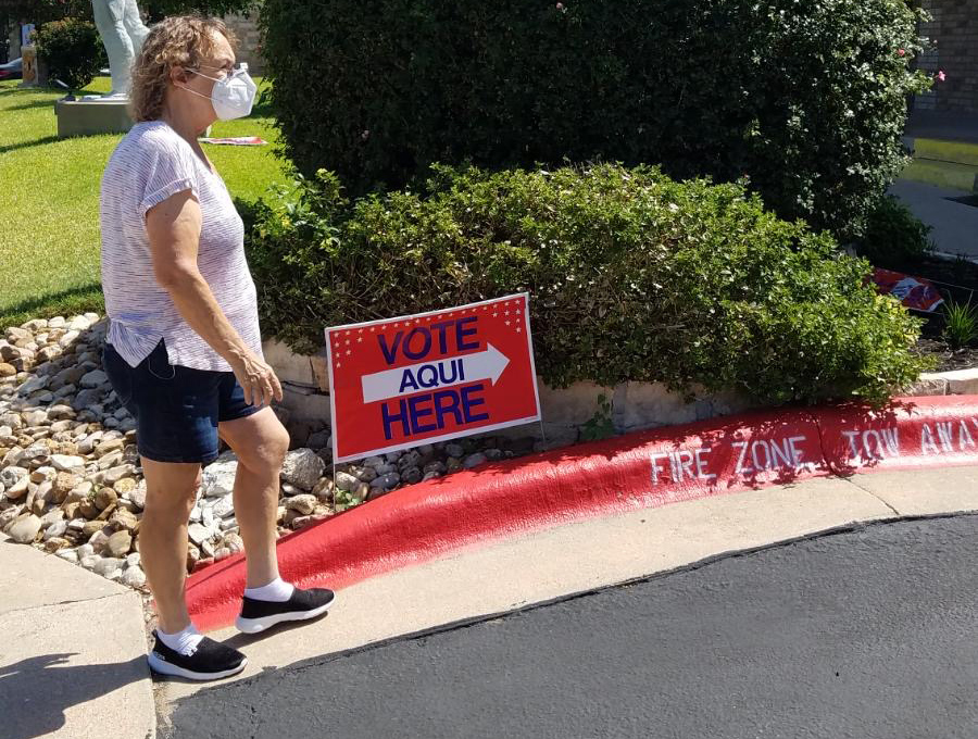 A White woman wearing a mask walks towards a polling place in a residential neighborhood. A sign reads “VOTE” with an arrow pointing in the direction she is walking.