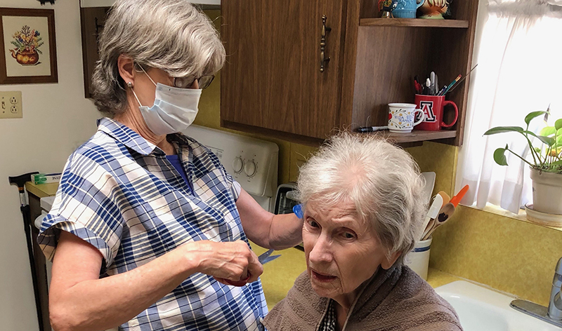 A middle-aged White woman wearing a mask cuts an older White woman’s hair at home.