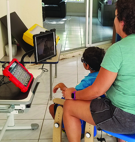 A school-age Multiracial boy sits in an adapted chair with a female adult sitting behind him. They are joined in an online meeting. An augmentative communication device sits to the left of the boy. 