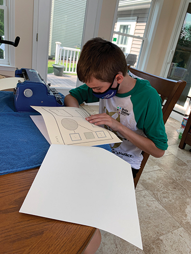 A young, White boy wearing a mask sits at a desk with a brailler and reads a brailled document.