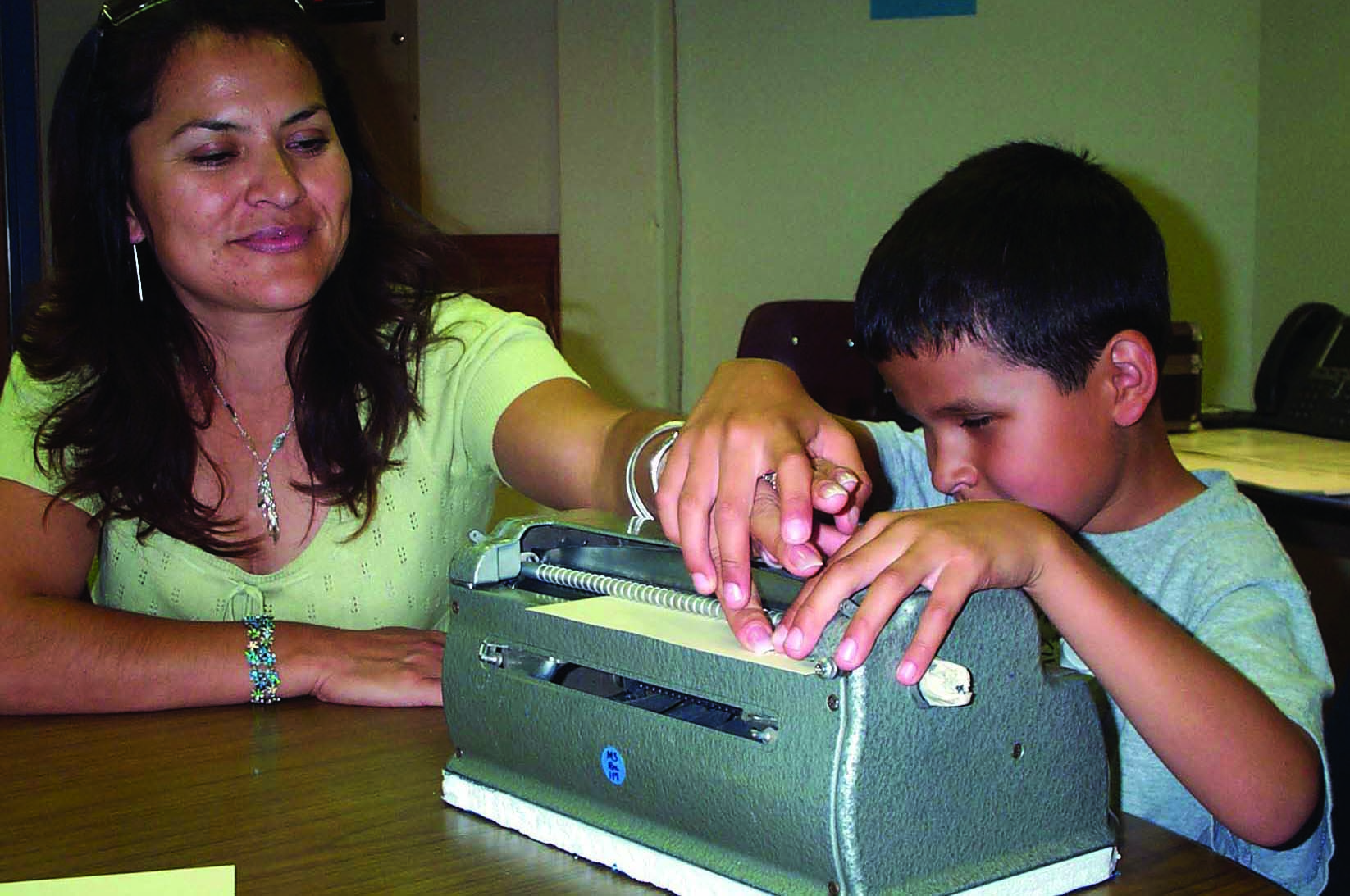 A Hispanic school-age boy takes his mother's hand to show her what he has brailled on his Perkins braillewriter. 