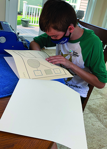 A White school-age teenage boy wearing a mask examines a tactile graphic of shapes prepared on microcapsule paper. 