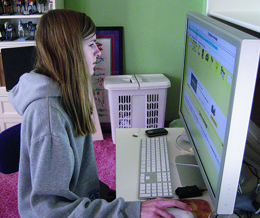 A White teenage girl looks at a large computer screen in her bedroom. 