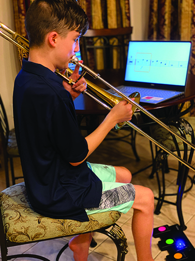 A White teenage boy sits on a chair playing his trombone. The music is magnified on a screen using the Lime Lighter from Dancing Dots. He controls the presentation of the music using the wireless pedal board that sits on the floor at his feet. 