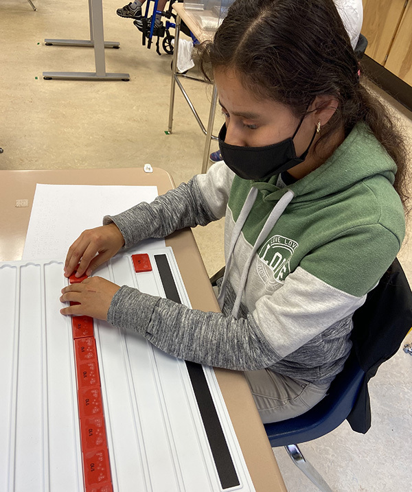 Young female student at her desk, wearing a mask, working with braille.