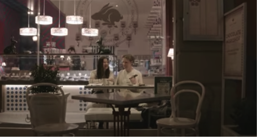 A still from a video showing two White people sitting next to each other in a restaurant, one holding a smartphone.