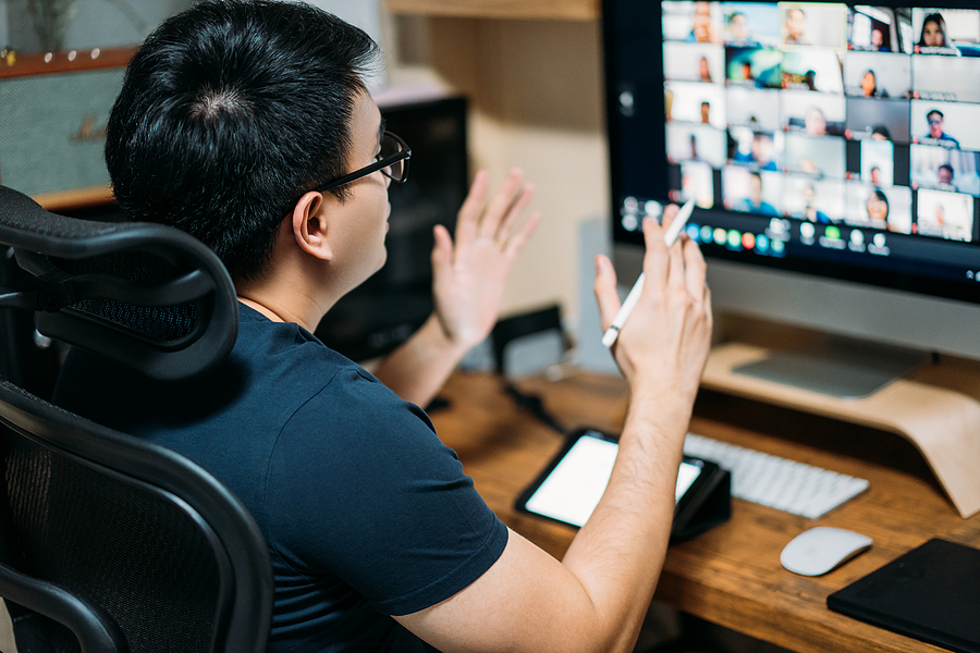 An Asian man sitting in his home office. He is using several pieces of technology and participating in a video conference.