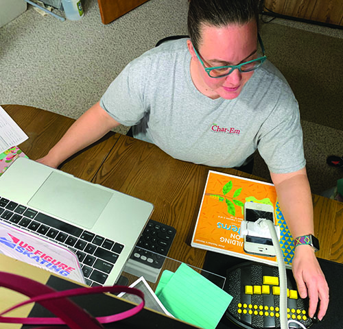 A White TVI in her home office has multiple devices set up on her desk including a laptop raised with a second keyboard underneath it. To her left a cell phone is attached to a gooseneck stand pointed at an APH Braille Bug while a copy of APH's Building on Patterns sits on the desk in front of her. 