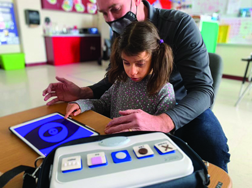 A White elementary school-aged girl uses an iPad with the app "Big Bang Pattern." Her White TVI sits behind her with his hands positioned to lend assistance if needed. To her left a communication device with five choices is visible. 