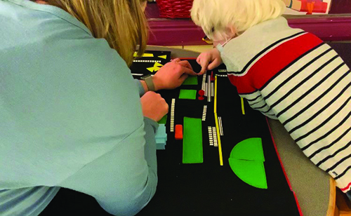 The photographer is looking over the shoulders of a White elementary boy wearing a mask and a White female O&M specialist as they explore a map created with APH's Tactile Town. 