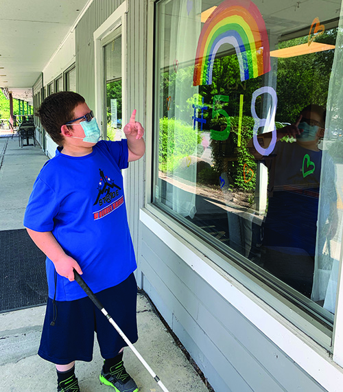 A White teenage boy wearing a mask points at an illustration on an outdoor window with his left hand and holds his cane in his right hand. 