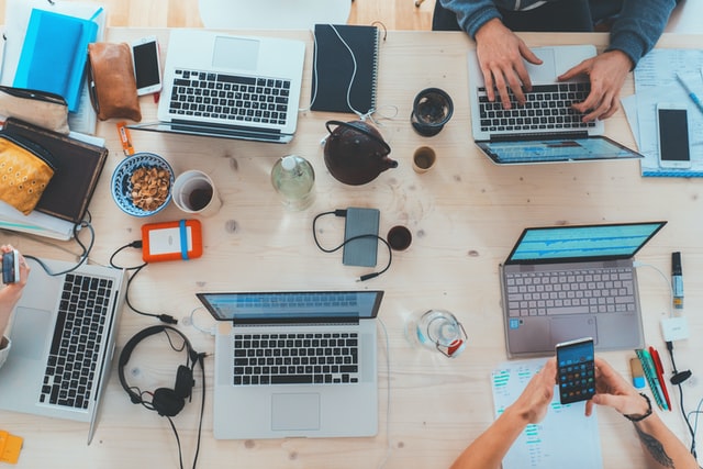 An overhead view of a table strewn with laptop computers, headphones, smartphones, notebooks, and other objects denoting “busy work.”