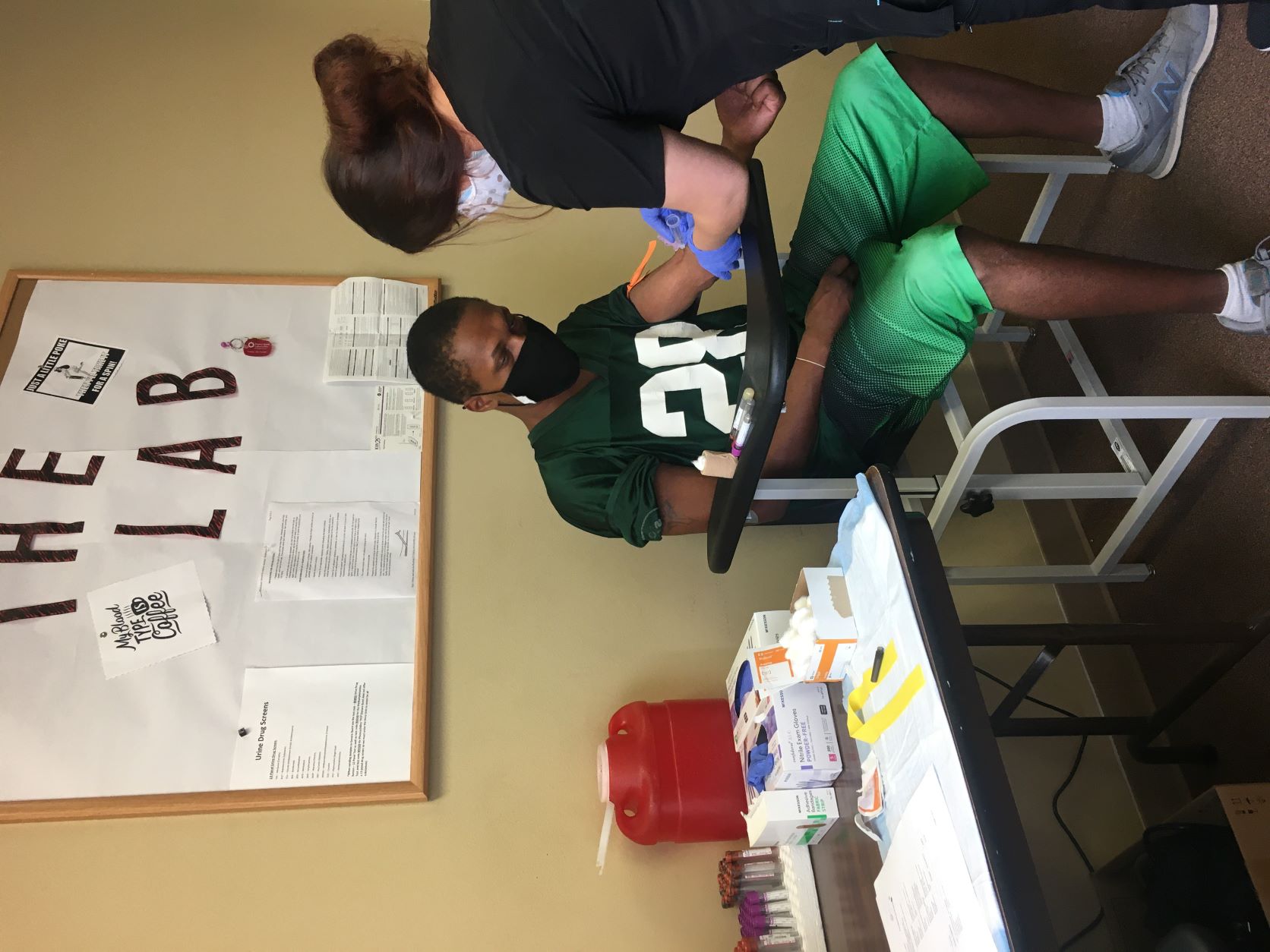  "A nurse draws blood from a young visually impaired African American man seated at a desk. Both are wearing masks."