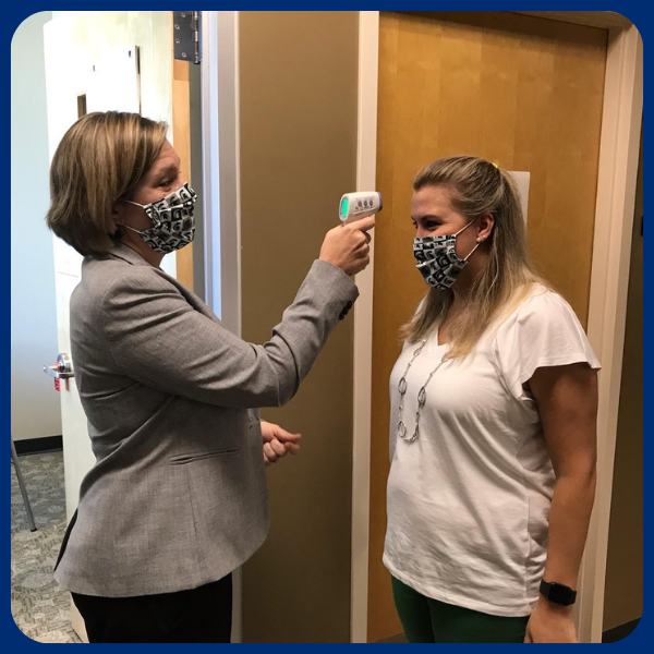 Two women, both wearing masks, stand at the entrance of a doorway. The woman on the left is using an infrared thermometer to take the temperature of the woman on the right.