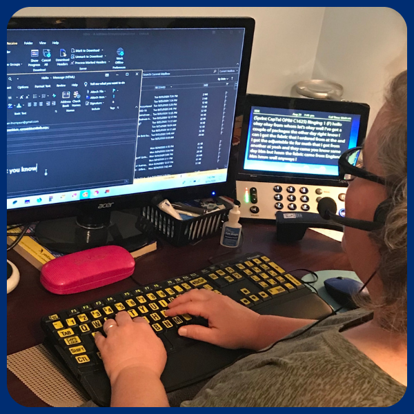 A woman sits at a desk in her home office. She is wearing a headset and typing on a computer keyboard with high-contrast keys while viewing her work on a computer monitor.