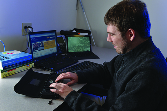 A man sits in front of two laptops and a braille notetaker