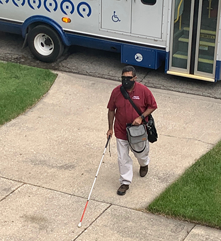 A man holding a white cane and wearing a mask has disembarked from a bus. He is walking away from the bus. The bus is in the background near the curb and has the passenger doors closed.