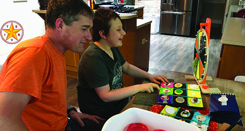 A White teenager sits at a kitchen table smiling with his hands resting on a game that has been adapted using tactile symbols. His father sits beside him and his instructor joins them online via an iPad resting on the table.