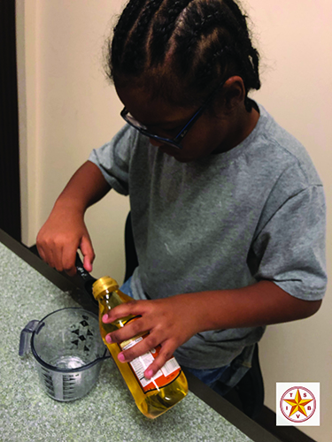 A Black school-age boy uses a measuring spoon to measures cooking oil into a measuring cup.