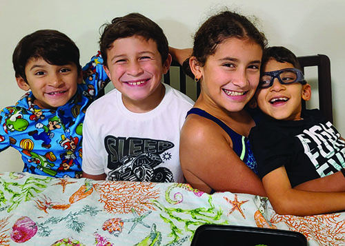 Three boys and a girl who are multiracial are celebrating a birthday. One boy sits on his sister's lap. He wears glasses and is missing his front teeth. All four siblings are smiling. 